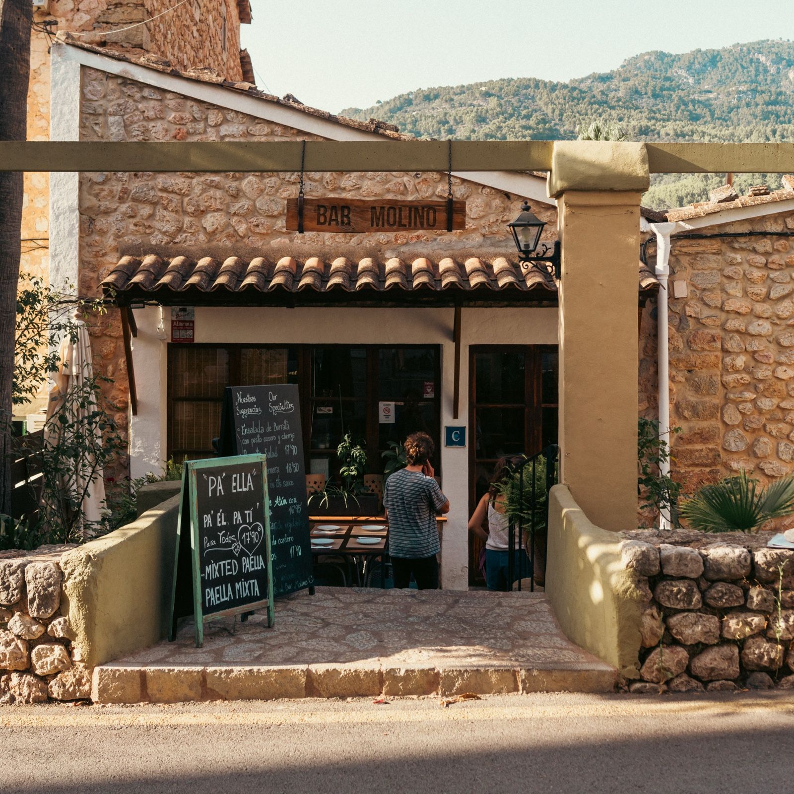 Fachada y entrada del Bar Molino en Sóller, con el cartel de madera, la pérgola y las montañas de fondo