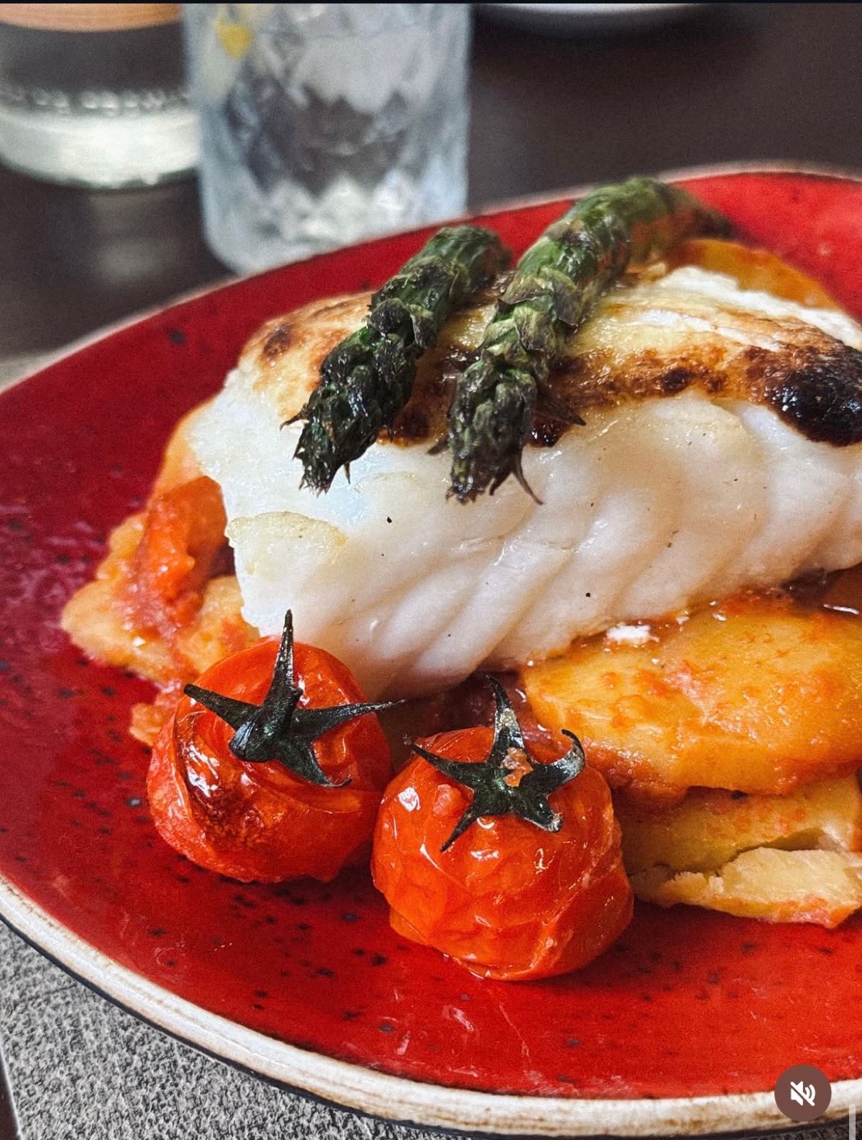 Oven-baked Jumbo cod with green asparagus, cherry tomatoes, a bed of tumbet (potato, aubergine and courgette) and gratinated aioli, on a red plate fresh from the oven.